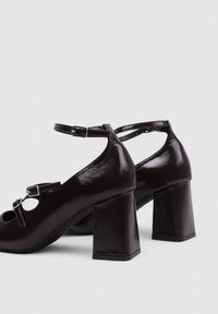 Escarpins en cuir verni marron foncé avec un large talon carré, une sangle autour de la cheville et des accentuations circulaires découpées au niveau des orteils. Fini brillant.