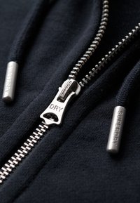 Sort zip-up hoodie med en sølvfarvet lynlås. Har fremtrædende metaldetaljer mærket "SUPERDRY" og teksturerede stofdetaljer.