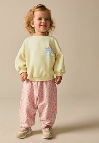 Sárga pulóver "hola" hímzéssel és kék hullámokkal, rózsaszín pöttyös nadrággal párosítva. Világos színű tornacipőkkel egészül ki az öltözék.