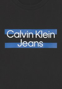 Camiseta de algodón negra con el texto blanco "Calvin Klein" superpuesto con barras azules, situado en el centro. Diseño clásico de cuello redondo.