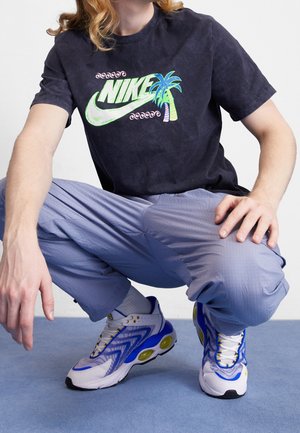 Camiseta negra con un vibrante logo de Nike y un gráfico de palmera. Pantalones texturizados de color azul claro. Zapatillas blancas con acentos en azul y amarillo.