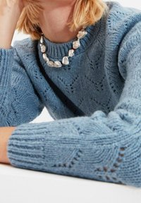 Hellblauer Strickpullover mit strukturierten Rautenmuster und gerippten Bündchen, kombiniert mit einer groben, unregelmäßigen Perlenkette.