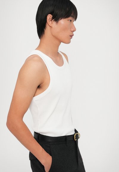 Top de tanque branco sem mangas com uma textura canelada, combinado com calças cinzas escuras e um cinto preto com fivela dourada. Vista lateral.