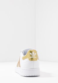 Bílé tenisky Adidas se zlatou patou a třemi zlatými pruhy, zobrazené zezadu na bílém povrchu a pozadí.