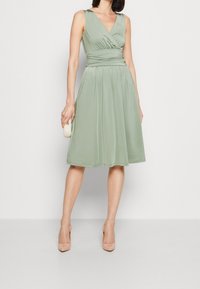 Salvia verde, vestido sin mangas con un cuerpo cruzado y cintura fruncida, fluye hacia una falda hasta la rodilla. Textura suave, combinada con tacones beig.