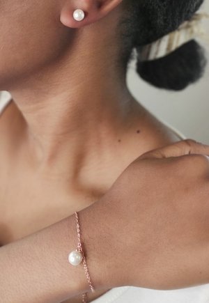 Boucles d'oreilles en perles et bracelet délicat en or rose avec accent en perle, sur un teint de peau neutre. Textures lisses et design simple.