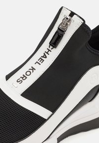 Sneaker nera slip-on con un upper in mesh testurizzato, dettagli in pelle bianca, una zip nera e stampa del logo sul lato.