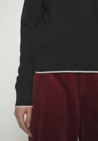 Pull en maille noir avec poignets côtelés et bordure blanche contrastante, associé à un pantalon en velours côtelé rouge avec des rayures verticales.