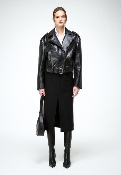 Žena koja nosi crnu kožnu motorističku jaknu, crnu midi suknju s prorezom sprijeda, crne čizme do koljena, bijelu košulju, drži crnu torbicu.