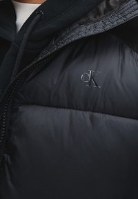 Černá péřová bunda s lesklým povrchem, se zapínáním na zip a embosovaným logem 'cK' na hrudi. Texturovaná látka v blízkosti límce.