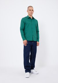 Grüne Hemdbluse mit langen Ärmeln, kombiniert mit dunkelblauen Jeans. Das Hemd hat einen klassischen Kragen und eine einfarbige Textur.