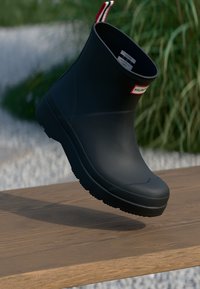 Cizmă din cauciuc neagră cu un design elegant, având un mâner cu dungi roșii și albe și o talpă texturată, expusă pe o suprafață de lemn.