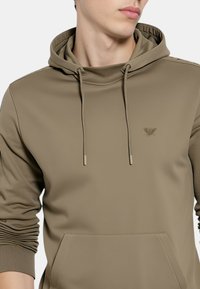 Felpa con cappuccio color kaki con coulisse, tasca a marsupio e polsini a costine. Presenta un piccolo logo in rilievo sul petto. Tessuto dalla texture morbida.