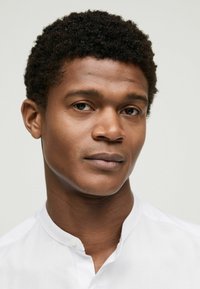 Hvid skjorte med krave uden knapper og med glat tekstur. Personen har kort, krøllet mørkt hår og et neutralt udtryk.