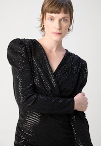 Top noir pailleté avec un design enroulé, un décolleté en v profond et des manches bouffantes. Les paillettes brillantes créent une surface texturée.