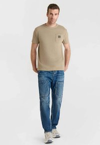 Maglietta a maniche corte color sabbia con una tasca, abbinata a jeans blu slavati con doppie tasche. Calzature sono sneakers grigio chiaro.