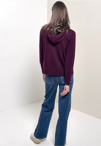 Lila Kapuzenpullover mit lockerer Passform, Kapuze und gerippten Manschetten, kombiniert mit blauen Schlagjeans und hellen Sneakers. Rückansicht.