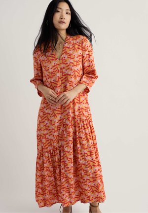 Frau mit langen schwarzen Haaren, die ein orangefarbenes, langärmeliges Maxikleid mit Blumenmuster trägt und vor einem schlichten hellen Hintergrund steht.