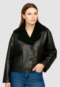 Giubbotto di pelle nera con ampio colletto, due tasche frontali e design corto, abbinato a jeans blu. Texture liscia, hardware minimale.