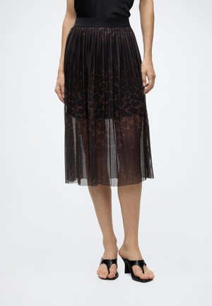Femme portant une jupe midi plissée noire avec un imprimé léopard foncé et une superposition transparente, associée à des sandales à talons ouvertes noires.
