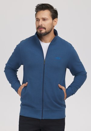 Mężczyzna w niebieskiej kurtce na zamek z rękami w kieszeniach, patrzy na bok na jednolitym jasnym tle.