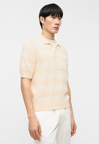 Πόλο μπλούζα σε ανοιχτό ροδακινί με κοντά μανίκια. Διαθέτει οριζόντιες υφασμάτινες ρίγες και γιακά που διπλώνει προς τα κάτω. Ριμπ τελείωμα στη μέση.