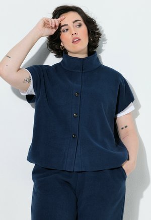 Donna con capelli corti e ricci che indossa un gilet blu navy abbottonato sopra una camicia bianca, con tatuaggi visibili su entrambi gli avambracci, posando contro un muro bianco.