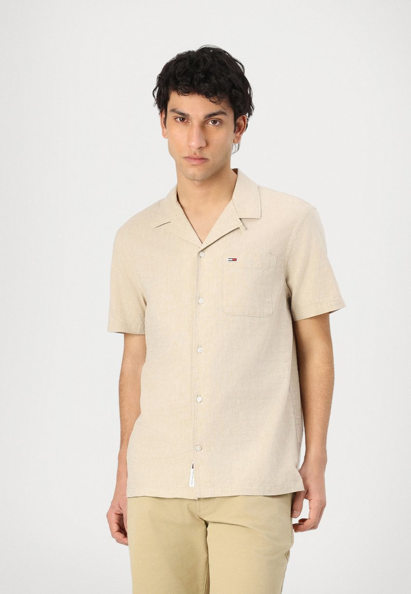 Kortermet beige skjorte med knapper og lomme, laget av teksturert stoff. Har en liten logo på brystet og en rett hem.