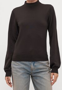 Person in einem dunkelbraunen Langarm-Mockneck-Pullover und hellblauen High-Waist-Jeans vor einem schlichten Hintergrund.