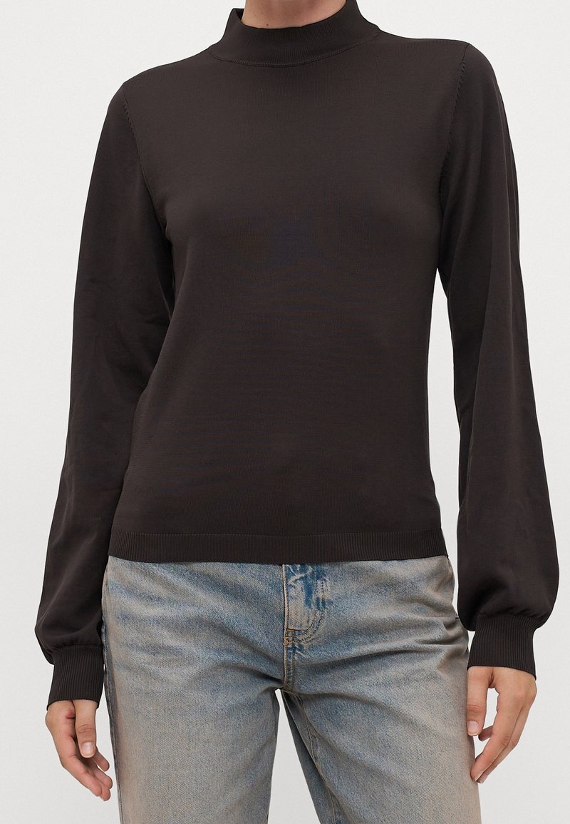 Person in einem dunkelbraunen Langarm-Mockneck-Pullover und hellblauen High-Waist-Jeans vor einem schlichten Hintergrund.