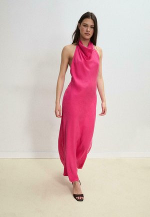 Ung kvinde går indendørs iført en ærmeløs lyserød halterkjole og sorte åbne hæle mod en ensfarvet baggrund.