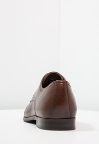 Bruna läderskor med en slät yta, elegant design, rund tå och en låg svart sula. Bakifrån framhävs sömnadsdetaljen.