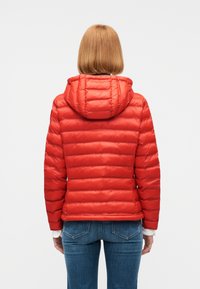 Άτομο με κοντά κόκκινα μαλλιά που φοράει κόκκινο καπιτονέ μπουφάν με κουκούλα και μπλε τζιν, με την πλάτη προς τα εμάς μπροστά σε απλό φόντο.