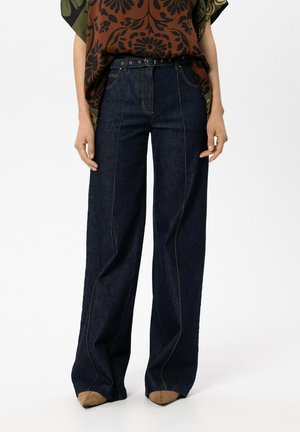 Person trägt hoch taillierte dunkelblaue Jeans mit weitem Bein, vorderen Nähten und Gürtel, kombiniert mit braunen spitzen Schuhen und einem Oberteil mit Blumenmuster.