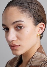 Boucle d'oreille en or en forme de croissant sur un modèle avec des cheveux plaqués en arrière, portant un blazer à motifs écossais. Teint neutre et maquillage naturel.