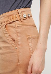Pantalons taille haute marron clair avec une finition texturée, dotés d'une poche latérale et de deux accents de boutons sur la ceinture.