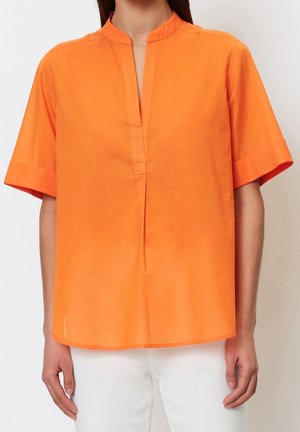 Personne portant un chemisier ample à manches courtes de couleur orange vif avec un col mandarin fendu, associé à un pantalon blanc.