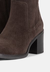 Stivaletti alla caviglia in suede marrone con punta rotonda e tacco a blocco nero. Presentano una texture liscia e dettagli di cucitura minimi.