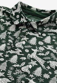 Tricou polo verde, cu un model alb de sărbători de Crăciun, având cerbi, copaci, stele și vâsc. Include un guler și un detaliu cu două butoane.