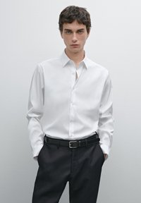 White long-sleeve shirt with a classic collar and button placket, paired with dark trousers and a black belt, against a neutral backdrop.