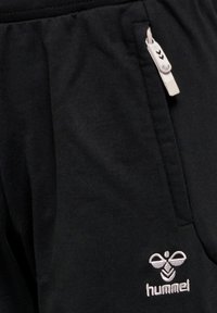 Svarta sweatpants med en slät textur, med en broderad vit logotyp längst ner till vänster och en beige dragsko i midjan.