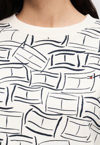 Λευκό πουκάμισο με στρογγυλή λαιμόκοψη, αφηρημένα μαύρα ορθογώνια σχέδια και μικρό κόκκινο, λευκό και μπλε λογότυπο στη δεξιά πλευρά του στήθους.