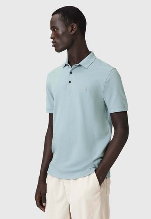Man die een lichtblauw polohemd met korte mouwen en beige broek draagt, staat met één hand in de zak, kijkt naar links tegen een effen achtergrond.