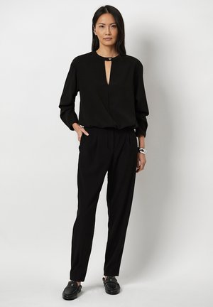 Zwarte blouse met lange mouwen en een kleine uitsnijding bij de hals, gecombineerd met zwarte op maat gemaakte broek en zwarte instappers met studs.