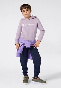 Fiú világoslila Champion kapucnis pulóverben, sötétkék melegítőnadrágban, olívazöld sportcipőben és derékra kötött lila pulóverben.