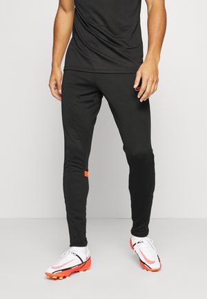 Man staat en draagt zwarte sportbroek met rood logo en wit-oranje voetbalschoenen op een lichtgrijze vloer en achtergrond.