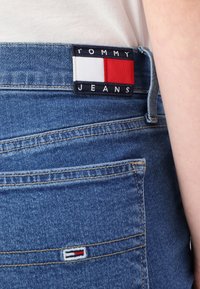 Jeans de mezclilla azul con una etiqueta azul marino que lleva el texto "TOMMY JEANS" y un diseño rectangular en rojo, blanco y azul en el bolsillo trasero.