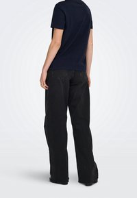 Personne portant une chemise bleu marine à manches courtes et un jean noir ample, debout de dos sur un fond clair.