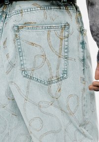Jeans din denim de culoare albastră deschisă, cu un model complicat de frânghie aurie, design cu cinci buzunare și accente cusute. Buzunarul din spate este vizibil.
