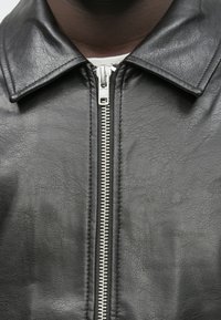 Casaco de pele preto com colarinho plano, fecho de correr diagonal e superfície texturada. Destaca-se o fecho de correr prateado ao longo do centro da frente.